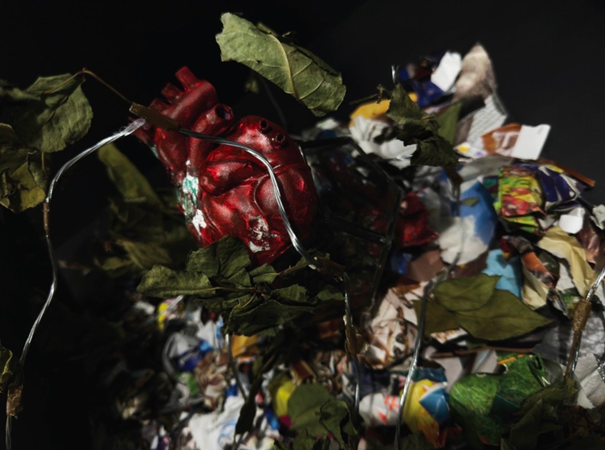 Tainted, detail: a red resin heart wound in wire among leaves and crumpled newsprint